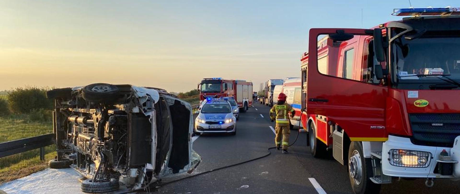 po lewej stronie przewrócony bus, przed nim poduszka z piany gaśniczej, po prawej samochód pożarniczy z rozwiniętą linią szybkiego natarcia, dalej radiowózy, karetka i samochód pożarniczy