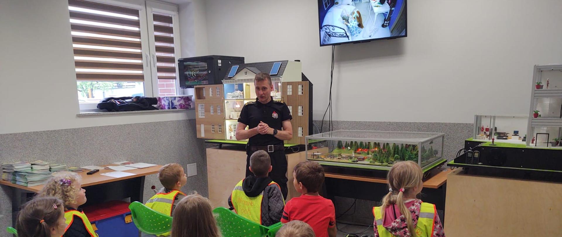 Na zdjęciu widać dzieci siedzące na krzesłach koloru zielonego. Przed dziećmi stoi strażak w umundurowaniu koszarowym koloru czarnego. Zdjęcie wykonane w pomieszczeniu. 