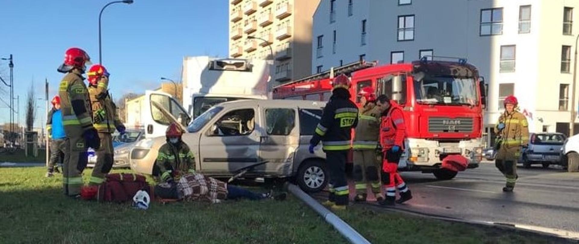Na zdjęciu widoczne są uszkodzone pojazdy, straż pożarna, pracujący strażacy, uszkodzona latarnia która leży na trawniku.
