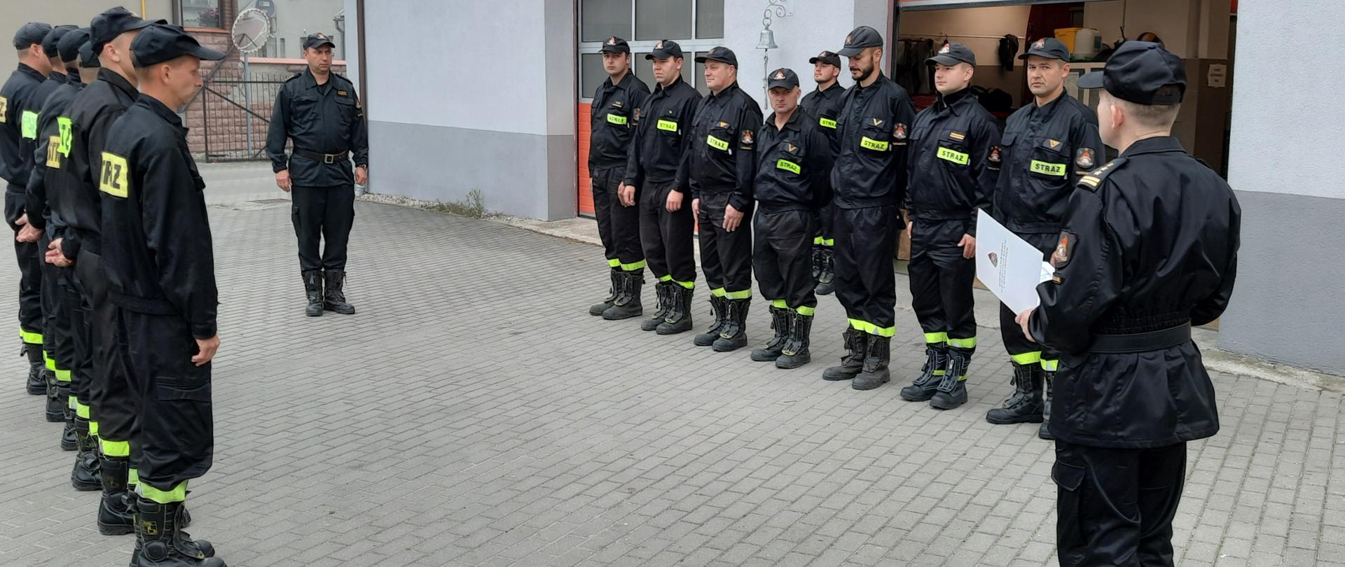 zdjęcie przedstawia strażaków Komendy Powiatowej PSP w Sępólnie Krajeńskim podczas zmiany służby. Komendant Powiatowy, który stoi bokiem odczytuje decyzję z okazji wręczenia nagrody jubileuszowej. Strażacy stoją - zmiana przyjmująca służbę po prawej stronie Komendanta, zmiana zdająca po lewej stronie Komendanta. Naprzeciwko Komendanta stoi zastępca dowódcy JRG. Wszyscy w umundurowaniu koszarowym. 
