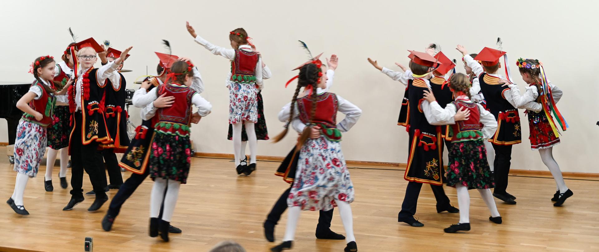 Uczniowie odziani w stroje krakowskie tańczą w parach na scenie sali koncertowej. Ogromna dynamika zdjęcia.