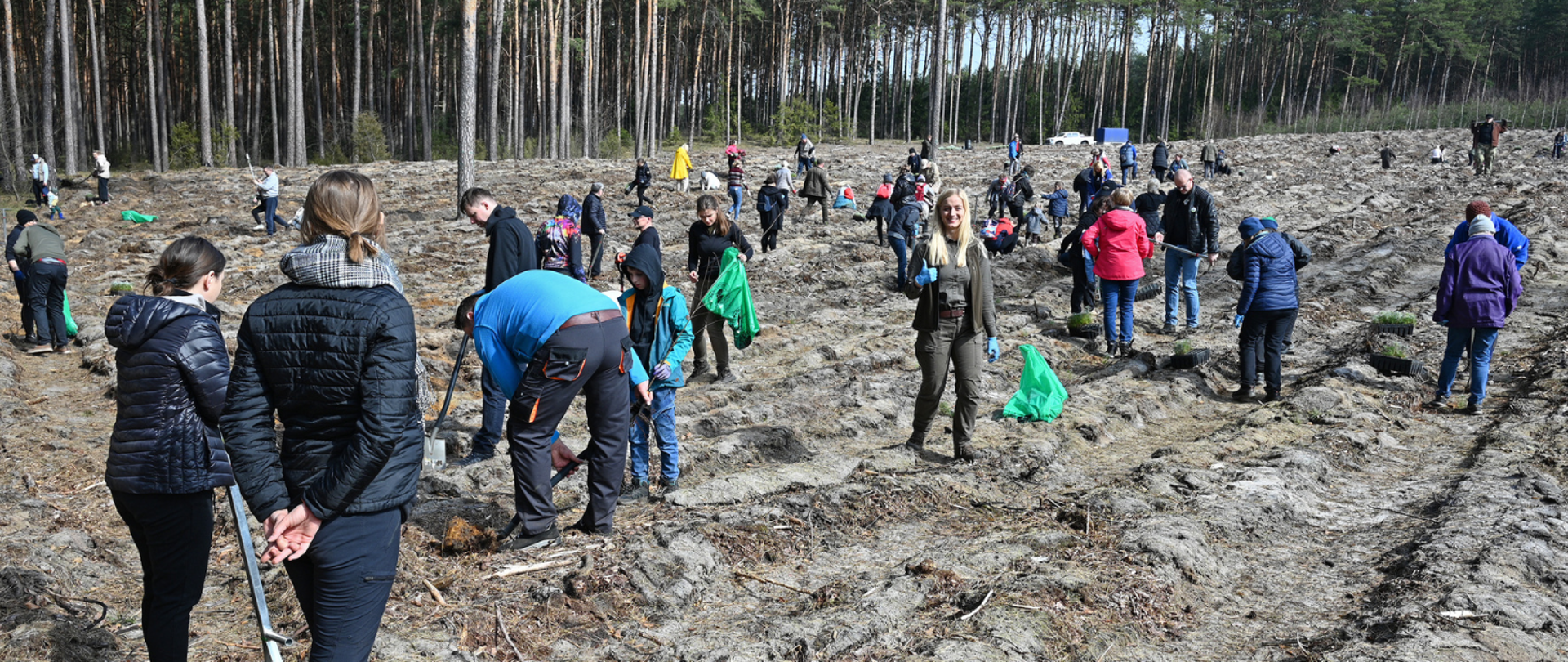 Grupa pracowników sadzi drzewa na otwartym terenie leśnym; uczestnicy pracują z łopatami i niosą sadzonki oraz worki.
