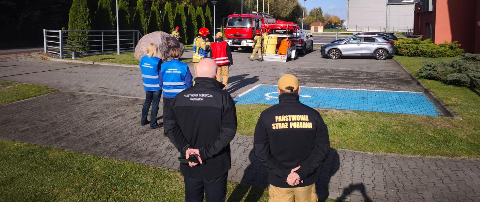 Fotografia przedstawia ćwiczenia służb mundurowych na placu przed budynkiem. Na pierwszym planie widoczne są sylwetki funkcjonariuszy Państwowej Straży Pożarnej oraz Państwowej Inspekcji Sanitarnej stojących tyłem do obiektywu. W tle znajdują się zaparkowane pojazdy ratownicze, w tym czerwony wóz strażacki, oraz grupa strażaków pracująca w pobliżu pamiątkowego głazu pod bezchmurnym niebem