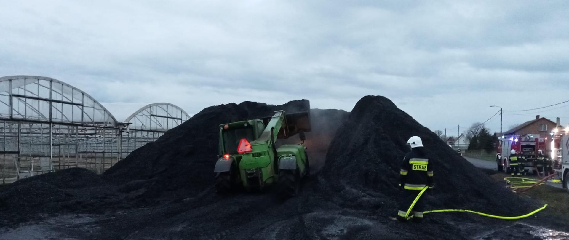 Na zdjęciu czarna hałda miału. Wjeżdża na nią zielona ładowarka. Obok strażak OSP w białym hełmie. Wąż fluo. Po prawej samochód pożarniczy.