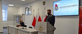 Kapelan wojewódzki strażaków przemawia podczas narady stojąc przy mównicy, przy stole siedzi pomorski komendant wojewódzki Państwowej Straży Pożarnej.