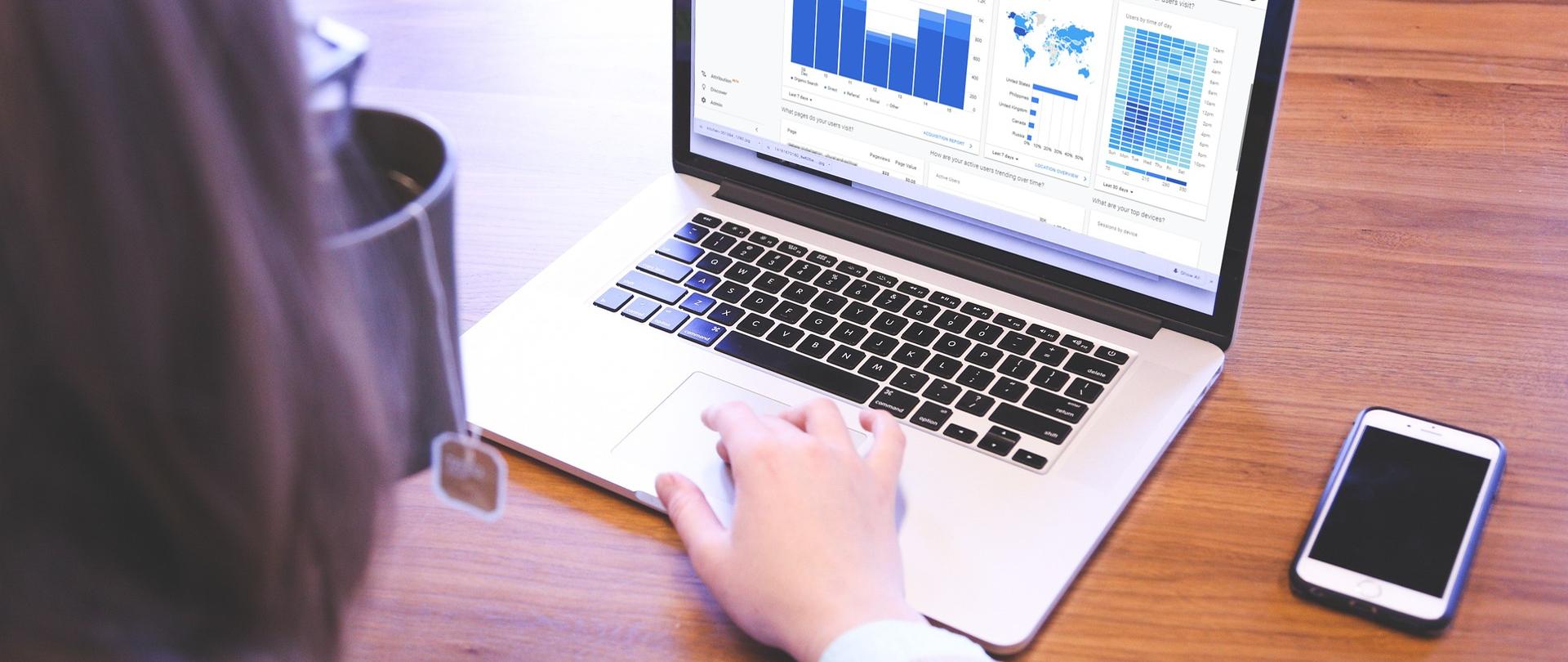 Na zdjęciu kobieta przegląda stronę internetową z wykresami na otwartym laptopie. Na stole obok laptopa leży telefon.