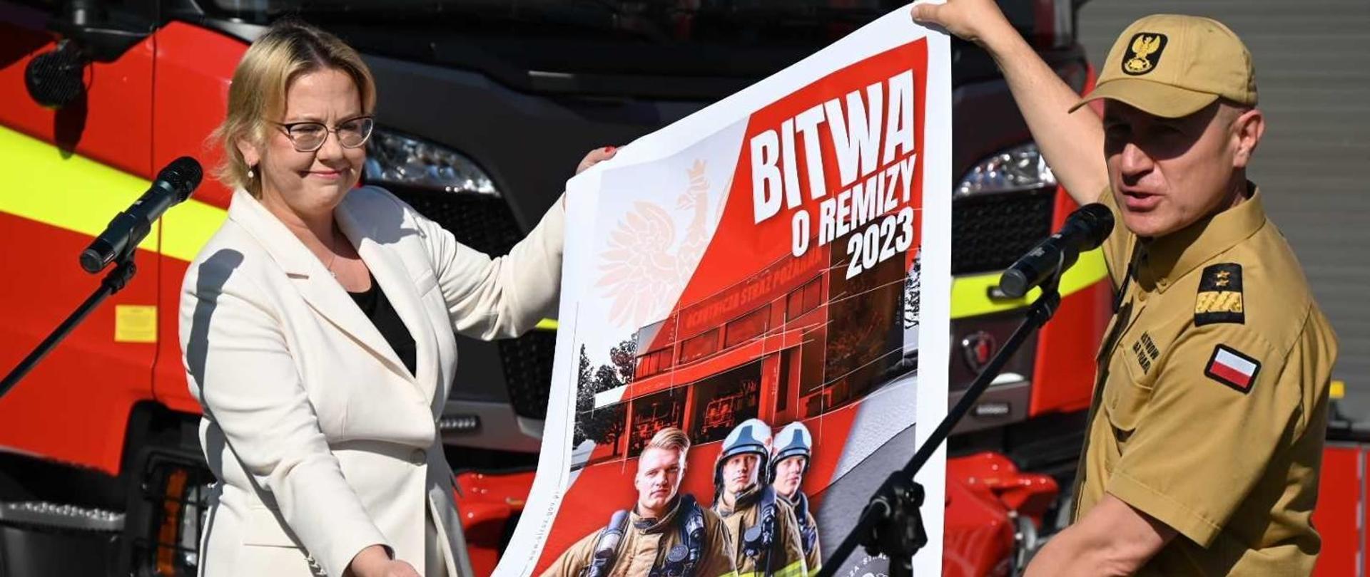  Komendant główny PSP trzyma plakat z panią minister „Bitwa o remizy” , w tle samochód strażacki.