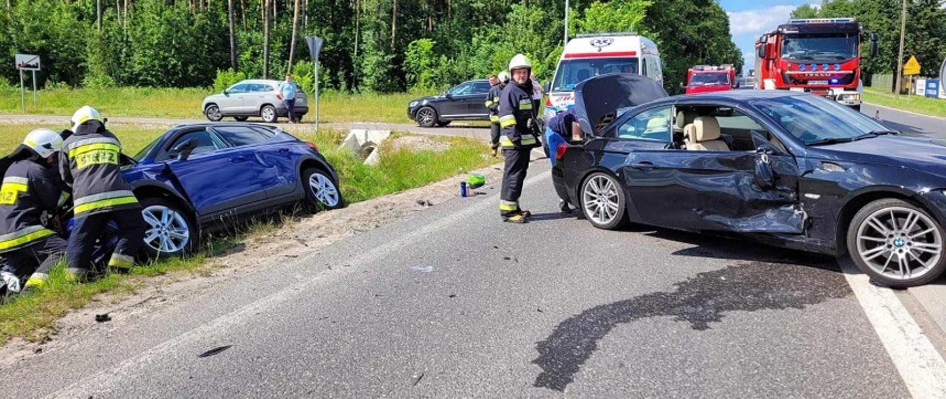 Z lewej strony znajduje się samochód Seat, który w wyniku zderzenia z samochodem BMW wpadł do rowu. Przed nim dwóch strażaków z Ochotniczej Straży Pożarnej rozłącza akumulator. Z prawej strony widzimy samochód BMW, który ma uszkodzony bok od strony pasażera. Z samochodu na ulicę wyciekły płyny eksploatacyjne. W tle zdjęcia znajduje się karetka pogotowia oraz pożarnicze samochody.