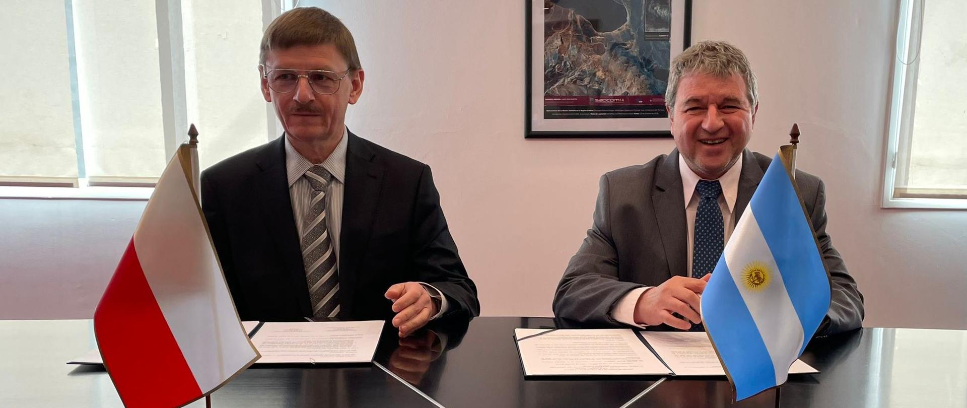 Prezes Polsy Grzegorz Wrochna i przedstawiciel argentyńskiej agencji kosmicznej siedzą przy stole i podpisują porozumienie o współpracy.
