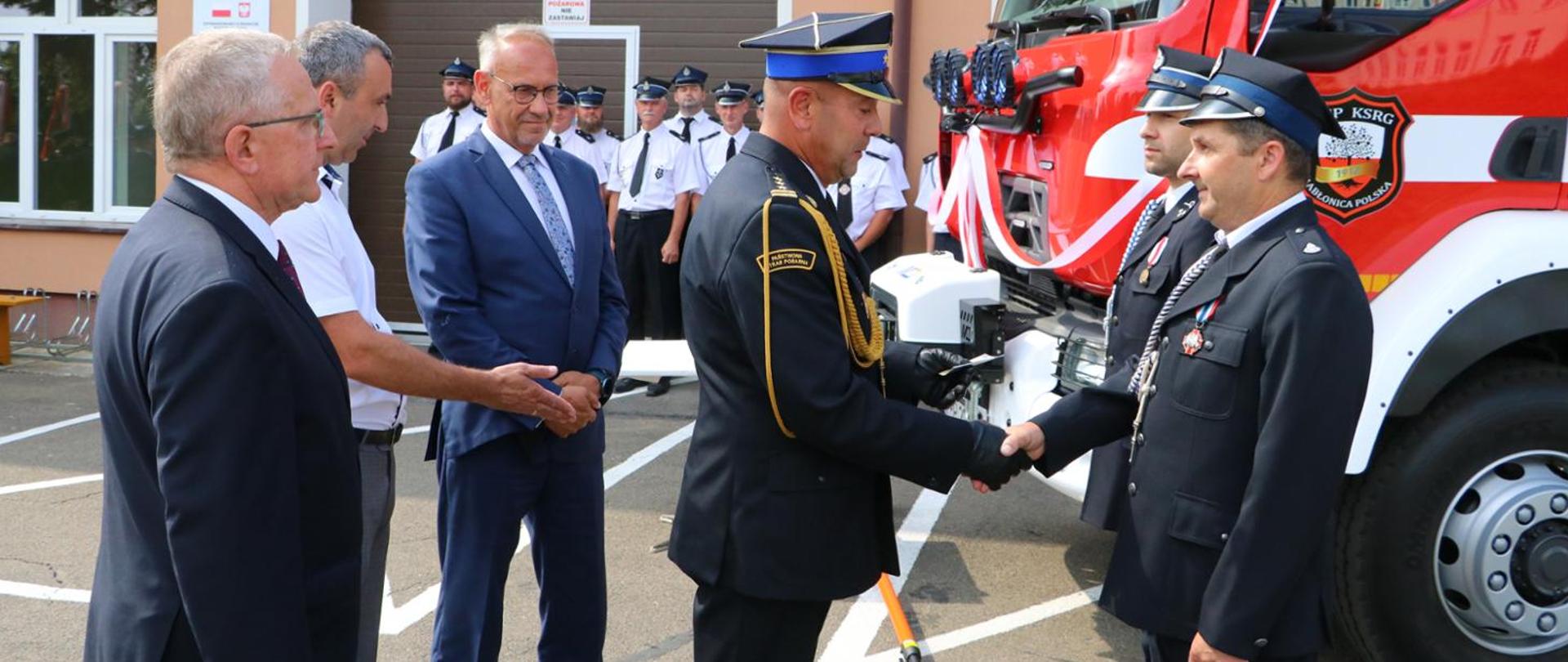 Kolorowa fotografia wykonana na zewnątrz w pogodny dzień. Przedstawia strażaka ubranego w mundurowaniu galowym PSP wręczający odznaczenia strażakom OSP wraz osobami cywilnymi. W tle nowy samochód pożarniczy przygotowany do poświęcenia.
