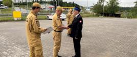 Na zdjęciu widać Komendanta Miejskiego PSP w Gdańsku wręczającego ubranemu w mundur wyjściowy PSP strażakowi, decyzję o nadaniu stanowiska Zastępcy Dowódcy Jednostki Ratowniczo-Gaśniczej nr 6. Jednocześnie Komendant uściskuje dłoń funkcjonariusza. Obok po lewej stronie, plecami do fotografa stoi Dowódca JRG nr 6, trzymający kartkę. W tle widać samochód służbowy Komendanta.