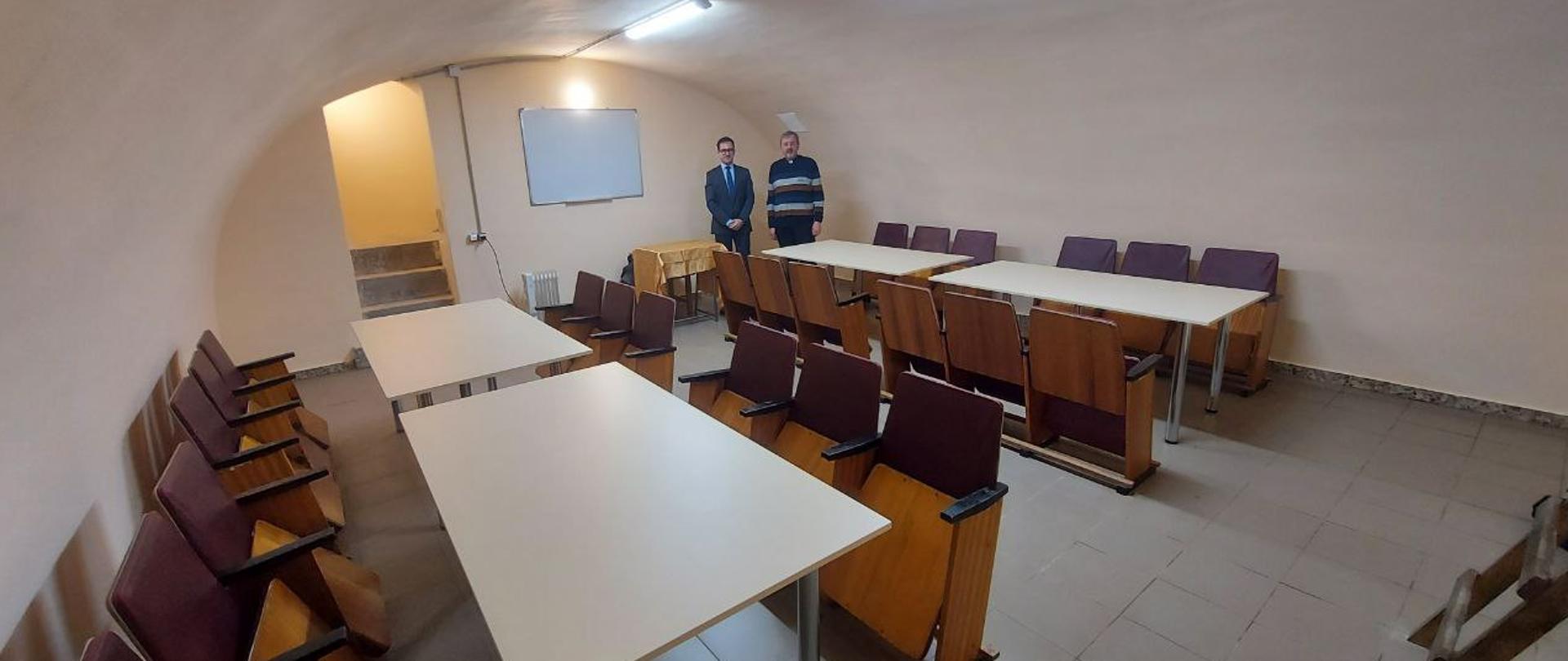 Schron, w którym znajdują się 4 stoły z krzesłami oraz tablica. Na końcu schronu stoi dwóch mężczyzn