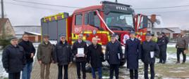 W rzędzie stoją osoby zaangażowane w pozyskanie środków na nowy samochód ratowniczo-gaśniczy, w tle nowe wóz strażacki.