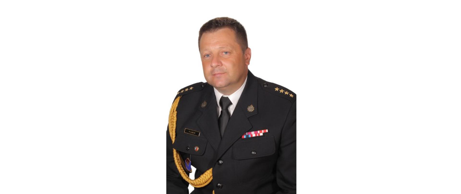 Mężczyzna w galowym mundurze Państwowej Straży Pożarnej, z widocznymi odznaczeniami, złotym sznurem na ramieniu i pagonami świadczącymi o wysokim stopniu. Mężczyzna ma krótko ścięte, ciemne włosy i poważny, skupiony wyraz twarzy.