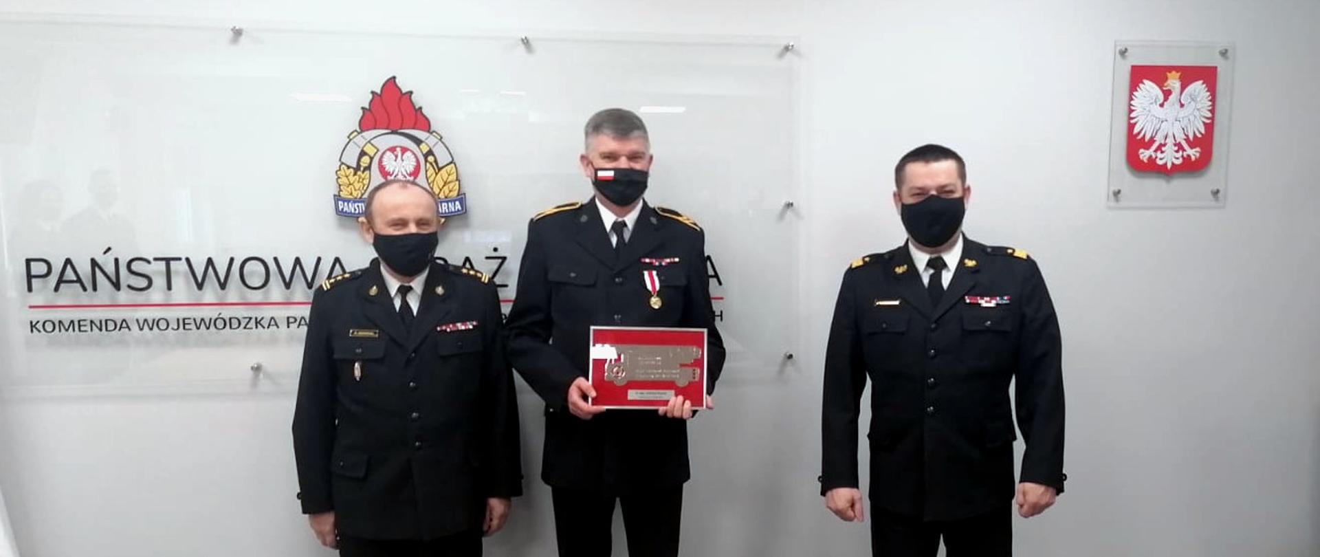 Zdjęcie przedstawia uroczyste pożegnanie odchodzącego na zaopatrzenie emerytalne st. ogn. Andrzeja Śmiecha. Na zdjęciu stoją w mundurach wyjściowych i maseczkach ochronnych: po prawej stronie Śląski Komendant Wojewódzki PSP - nadbryg. Jacek Kleszczewski, po lewej Zastępca Śląskiego Komendanta Wojewódzkiego PSP - st. bryg. Bogdan Jędrocha, w środku st. ogn. Andrzej Śmiech, który w ręce trzyma pamiątkową tabliczkę. W tle, na ścianie logo PSP, a po nim napis o treści Państwowa Straż Pożarna Komenda Wojewódzka PSP w Katowicach.