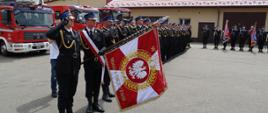 strażacy w mundurach galowych stoją w dwu szeregu, na pierwszym planie sztandar komendy 