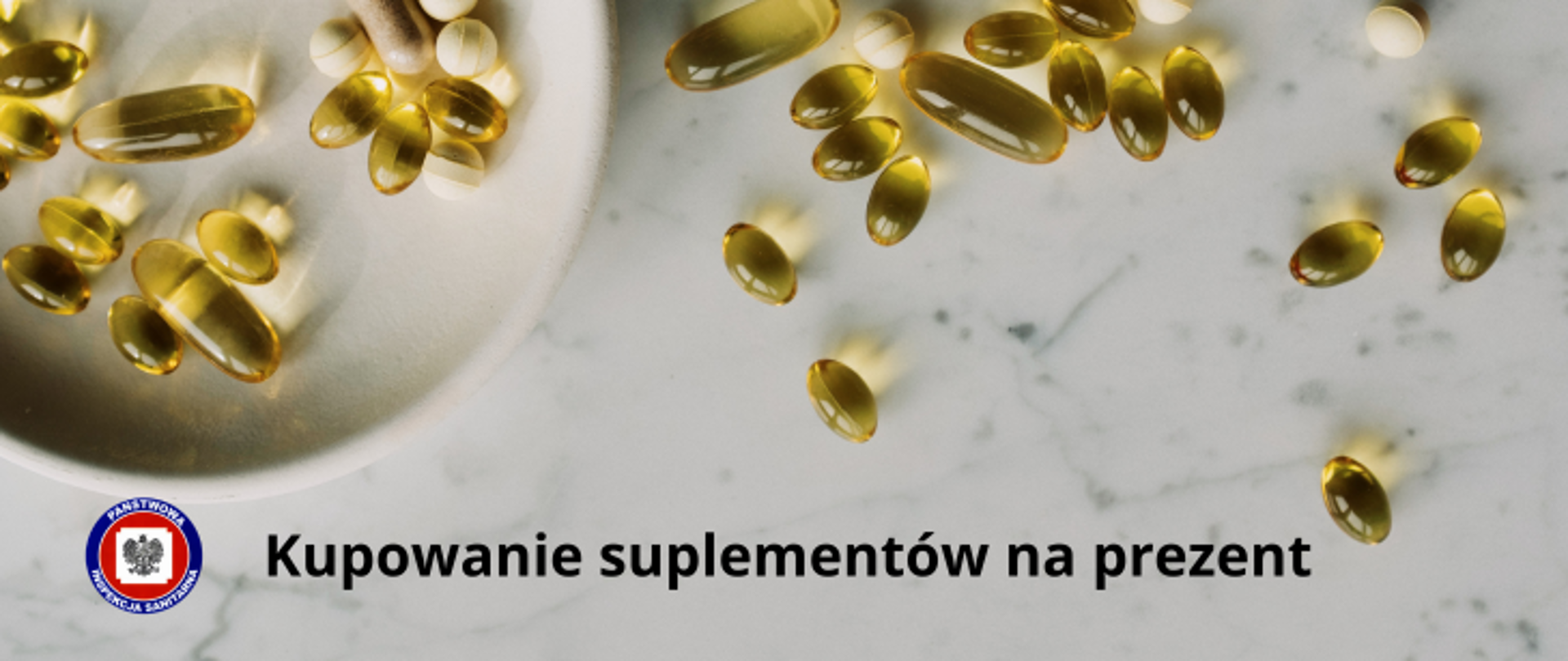 Na marmurowym stole leżą rozsypane różne tabletki z suplementami, z lewej strony stoi talerz jasnego koloru również zawierający różnego rodzaju tabletki. Poniżej czarny napis Kupowanie suplementów na prezent a z jego lewej strony logo Państwowej Inspekcji Sanitarnej.