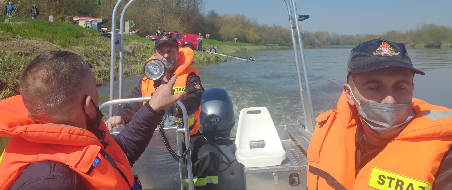 Zdjęcie przedstawia strażaków płynących łodzią, którzy przeszukują obszar wodny na rzece Wisła 