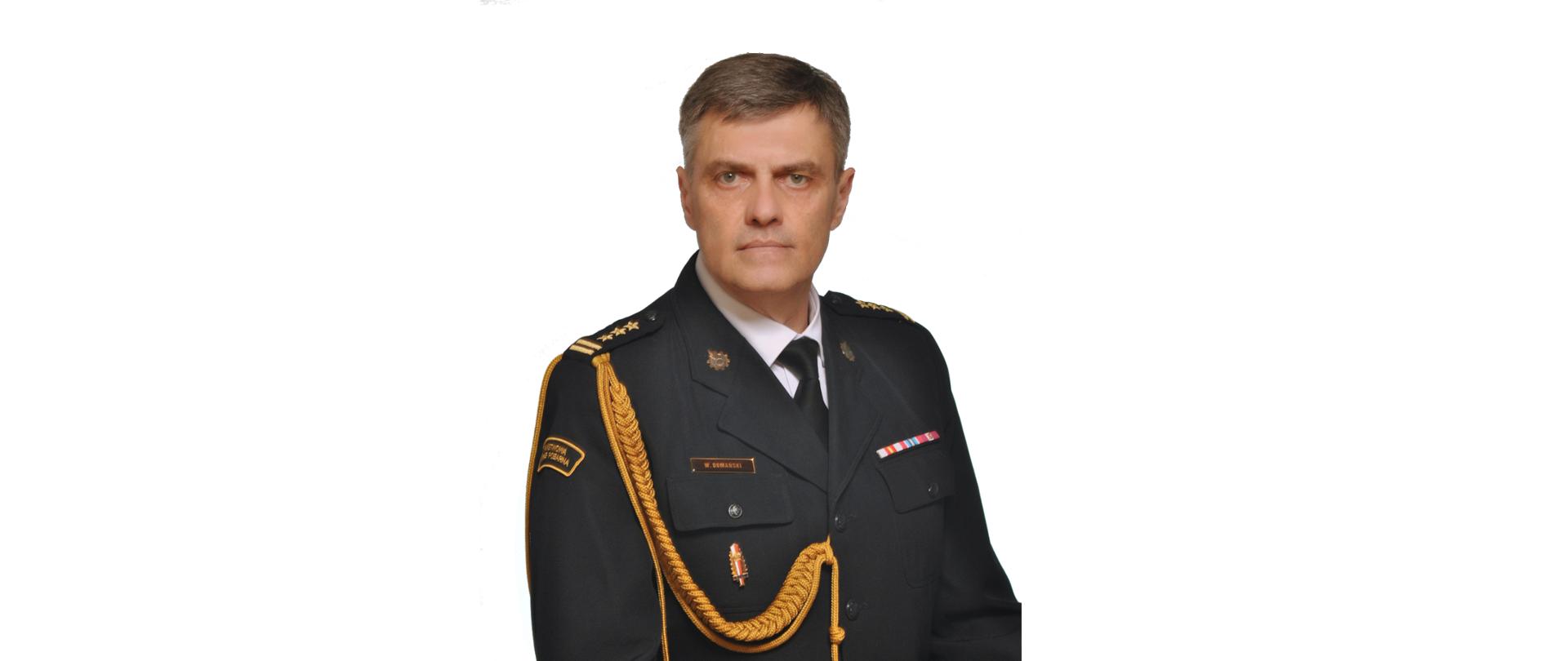 Portretowy, studyjny wizerunek mężczyzny w średnim wieku w ciemnym, galowym mundurze służbowym. Mężczyzna ma krótkie, siwiejące włosy i poważny wyraz twarzy. Na mundurze widoczne są odznaczenia, złote sznury ozdobne na ramieniu oraz pagony z oznaczeniami stopnia. Na piersi znajduje się imiennik z nazwiskiem. Na rękawie munduru widoczna jest naszywka z napisem w języku polskim. Tło zdjęcia jest jasne i neutralne