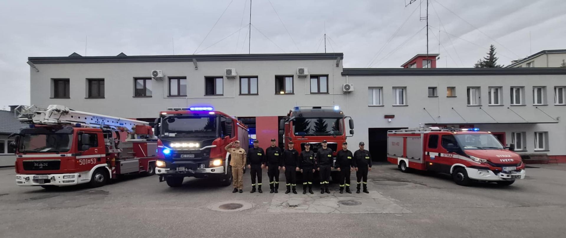 Strażacy z KP PSP w Makowie Mazowieckim oddają hołd zmarłemu druhowi. Z tyłu stoją cztery samochody strażackie a za nimi budynek komendy.