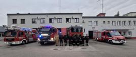 Strażacy z KP PSP w Makowie Mazowieckim oddają hołd zmarłemu druhowi. Z tyłu stoją cztery samochody strażackie a za nimi budynek komendy.