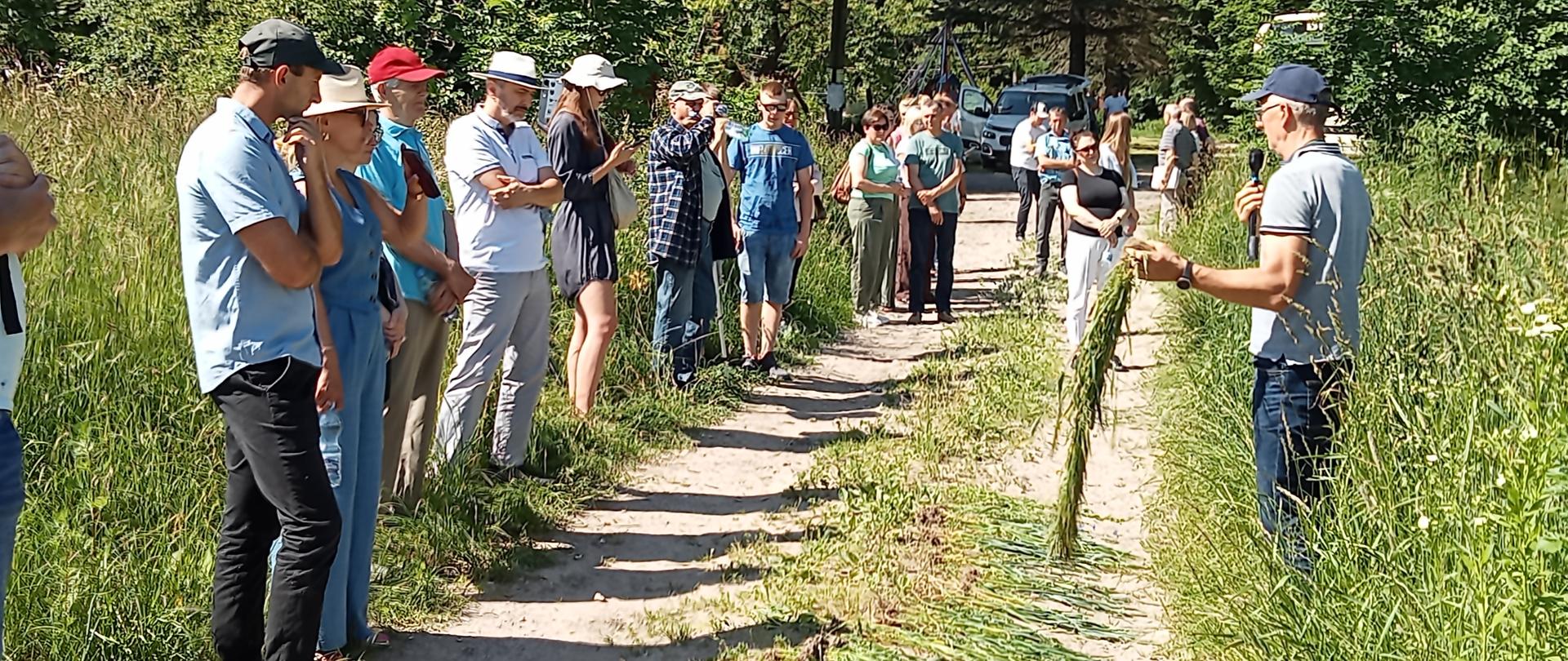 Mężczyzna stoi na skraju łanu zbóż, trzymając w rękach mikrofon i garść roślin, po drugiej stronie polnej drogi stoi grupa słuchających go osób