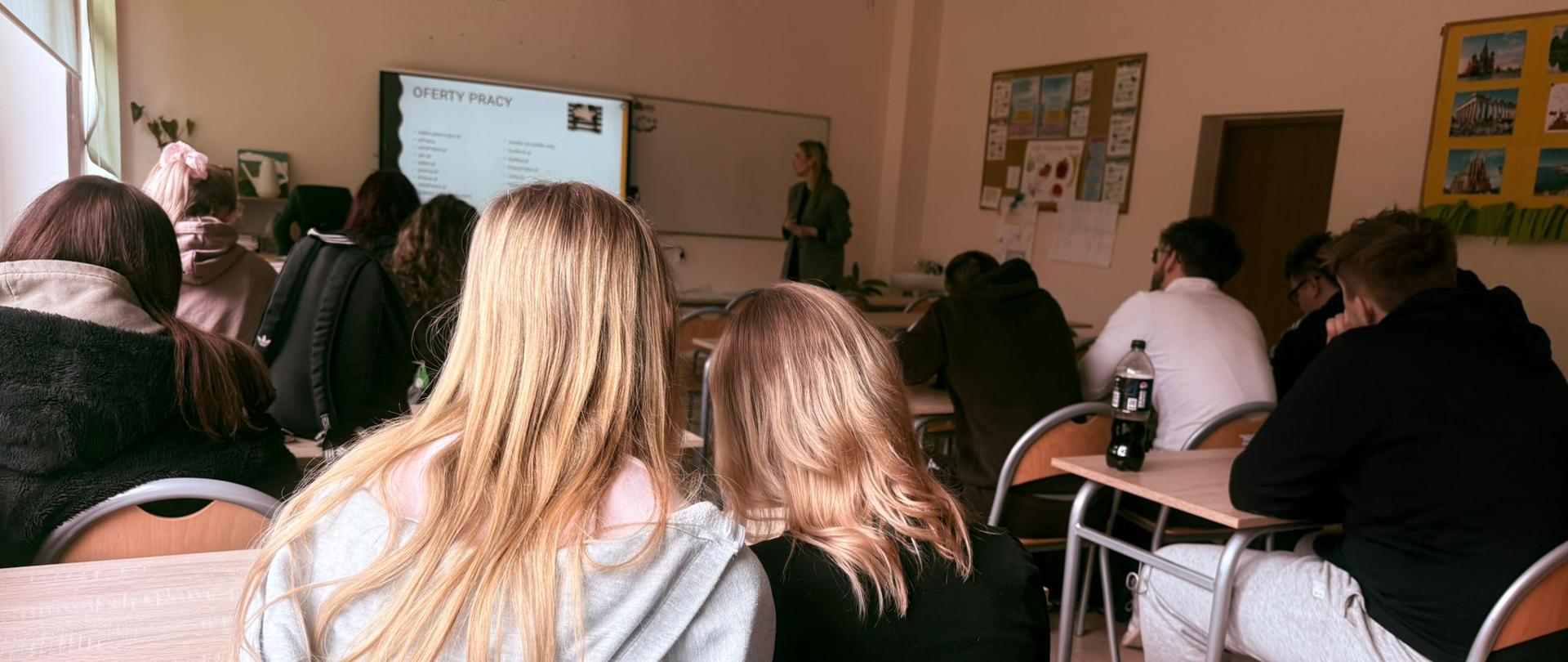 Widok zza pleców dwóch uczennic o jasnych włosach, siedzących w ławce lekcyjnej. W głębi sali, przy tablicy, stoi prelegentka, a obok niej ekran multimedialny z wyraźnym tytułem slajdu: „OFERTY PRACY”. Inni uczniowie siedzą w pozostałych ławkach.