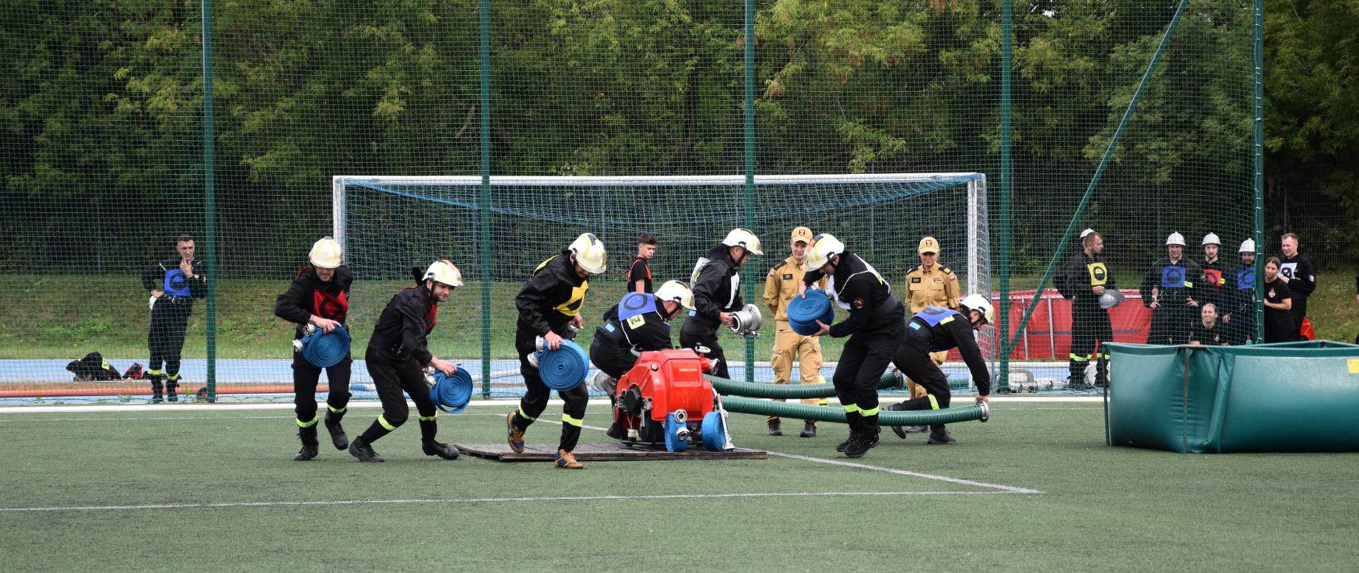 Strażacy podczas konkurencji ćwiczenia bojowego. Strażacy zabierają swój sprzęt z podestu. Część z nich idzie w stronę zbiornika z wodą z wężami ssawnymi, a część z nich zmierza biegiem w stronę linii mety ze swoimi wężami i pozostałym sprzętem. Za drużyną sędziowie przyglądają się wykonywaniu ćwiczenia. W tle za siatką boiska pozostali strażacy z innych drużyn.