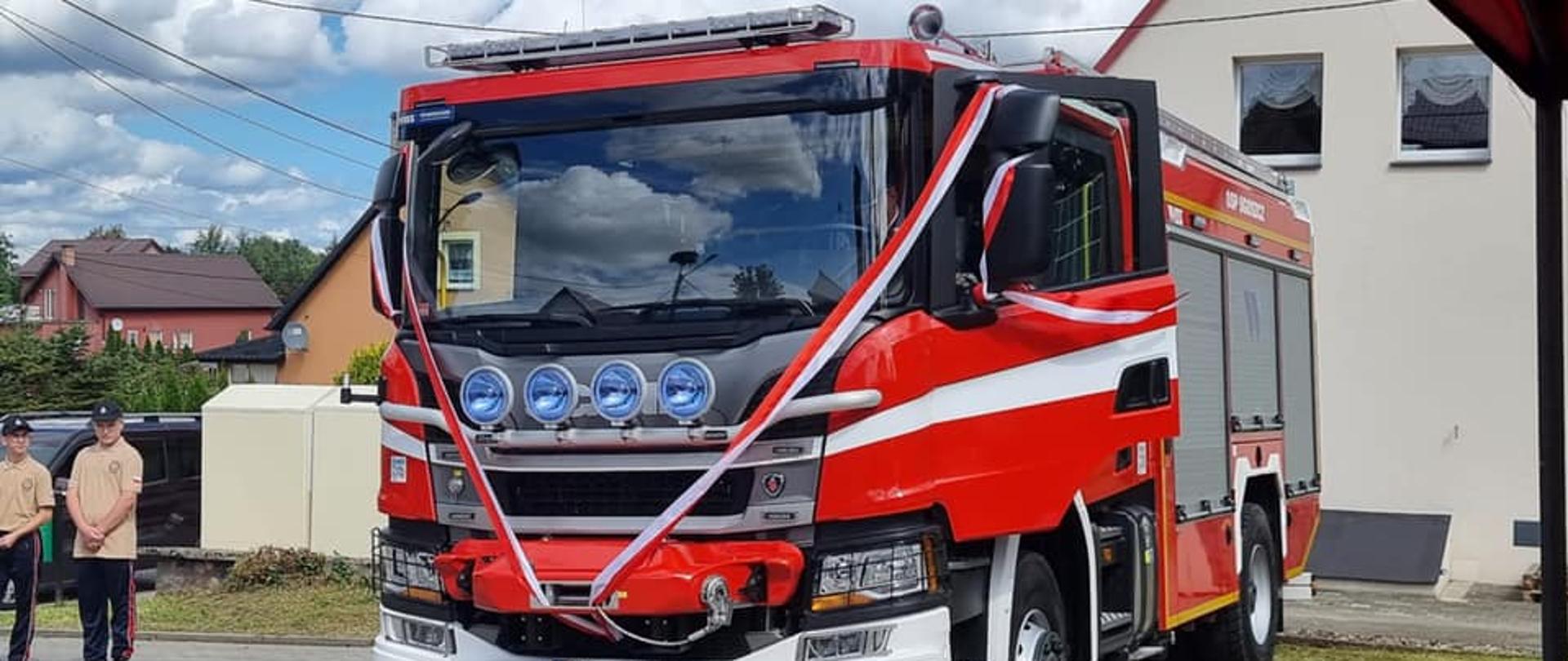Zdjęcie przedstawia ciężki samochód ratowniczo-gaśniczy koloru czerwonego, który jest udekorowany biało-czerwonymi wstążkami.