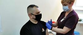 Strażak w umundurowaniu dowódczo sztabowym w czarnym podkoszulku przyjmuje szczepionkę