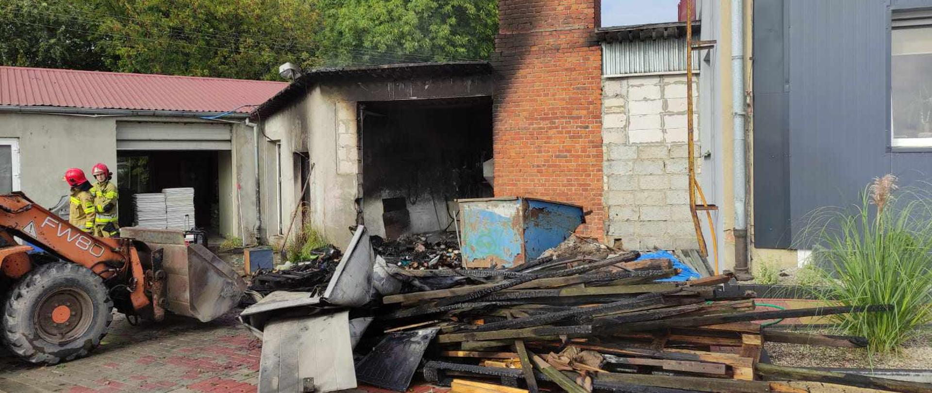 Zdjęcie przedstawia drewniane i spalone elementy wyniesione z kotłowni. Obok komin z cegły. Po lewej stronie stoi dwóch strażaków i koparka.