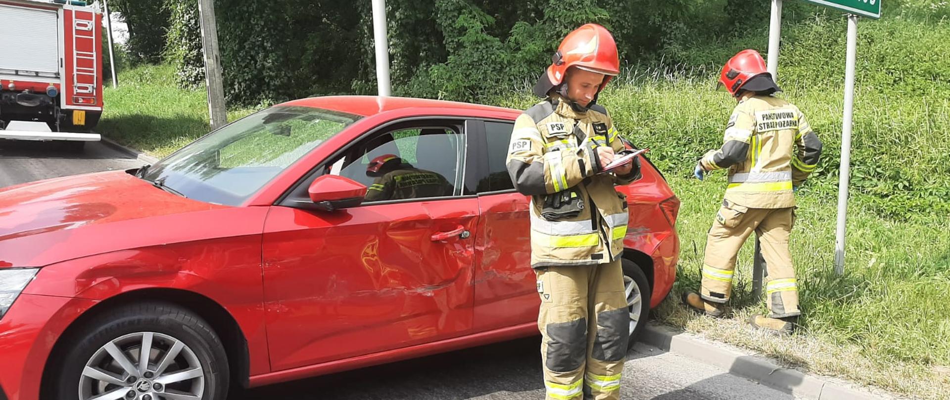 Czerwony samochód osobowy stoi w poprzek drogi. Przed nim strażacy, Jeden z nich poisze.