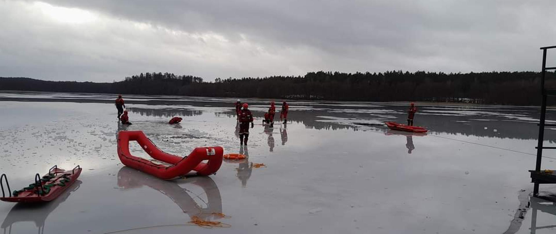 Strażacy w specjalistycznym umundurowaniu stoją na zamarzniętym jeziorze niedaleko jest specjalistyczny sprzęt sanie lodowe oraz pompowane sanie lodowe.