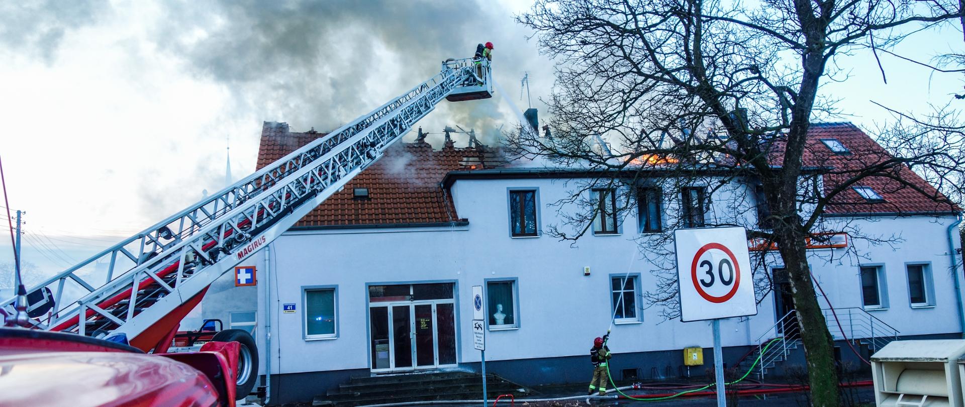 Strażacy w koszu drabiny mechanicznej podają prąd wody na palące się poddasze.