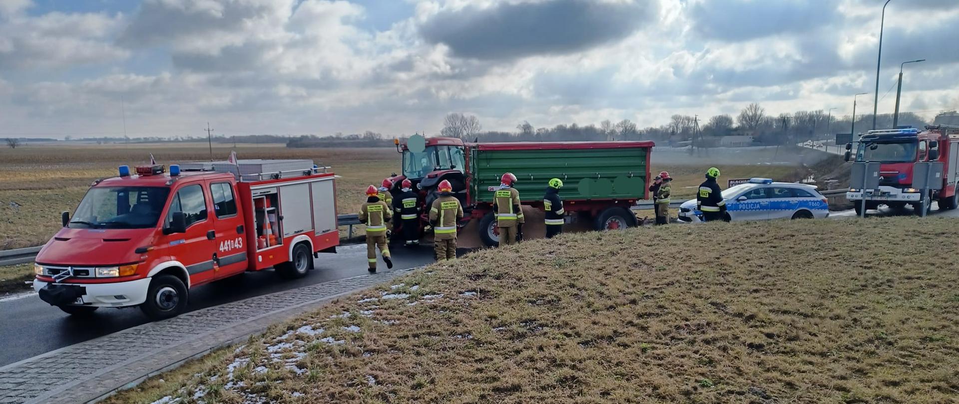 Na zdjęciu widać czerwony traktor z zieloną przyczepą, z której na jezdnie wysypało się zboże. Traktor stoi odwrócony tyłem do kierunku jazdy. Przed traktorem stoi czerwony samochód straży pożarnej, obok stoją strażacy w żółtych i czarnych strojach oraz żółtych i czerwonych hełmach. Za przyczepą stoi srebrny samochód policyjny a za nim czerwony samochód straży pożarnej. Całe zdarzenie jest na rondzie. W tle widać łąki oraz drzewa. Jest widno, ale pochmurnie.