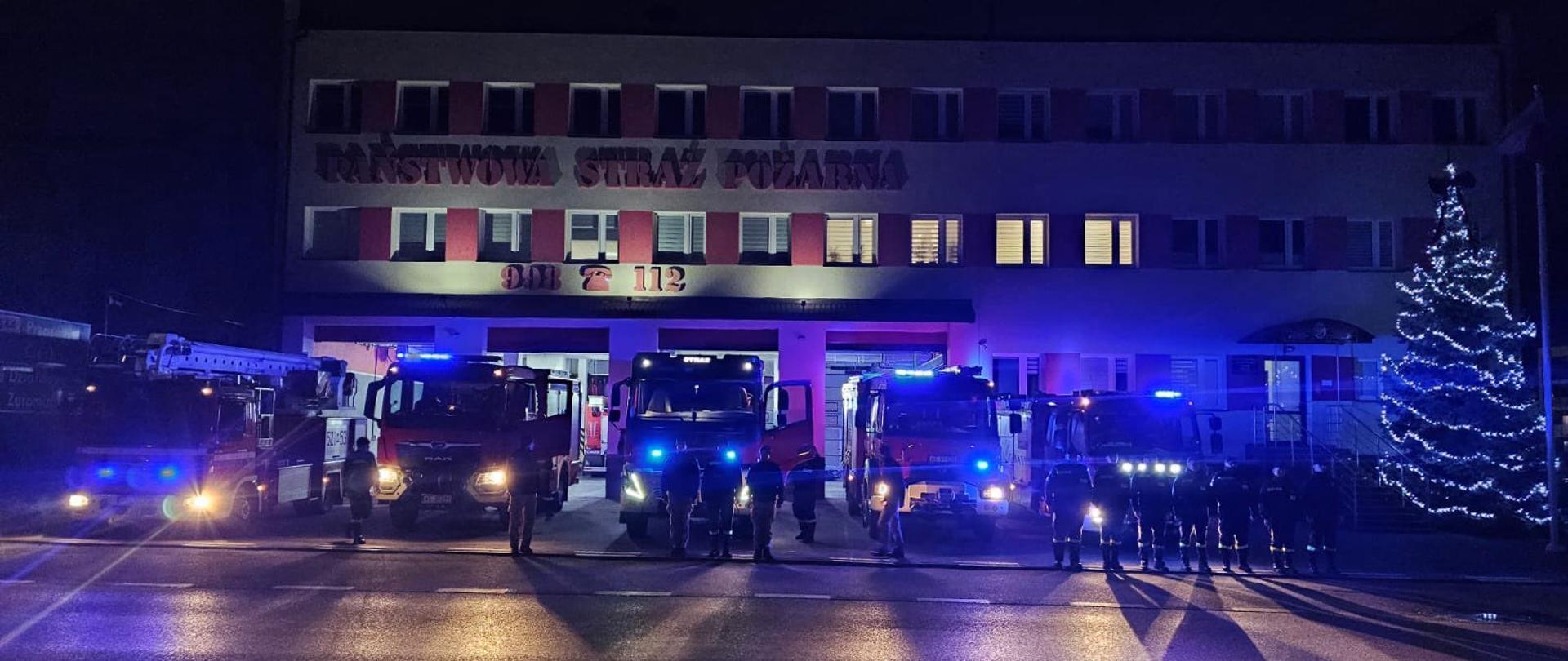 Przed budynkiem jednostki ratowniczo-gaśniczej stoją zaparkowane samochody strażackie z uruchomionymi niebieskimi światłami. Przed samochodami stoją strażacy PSP.