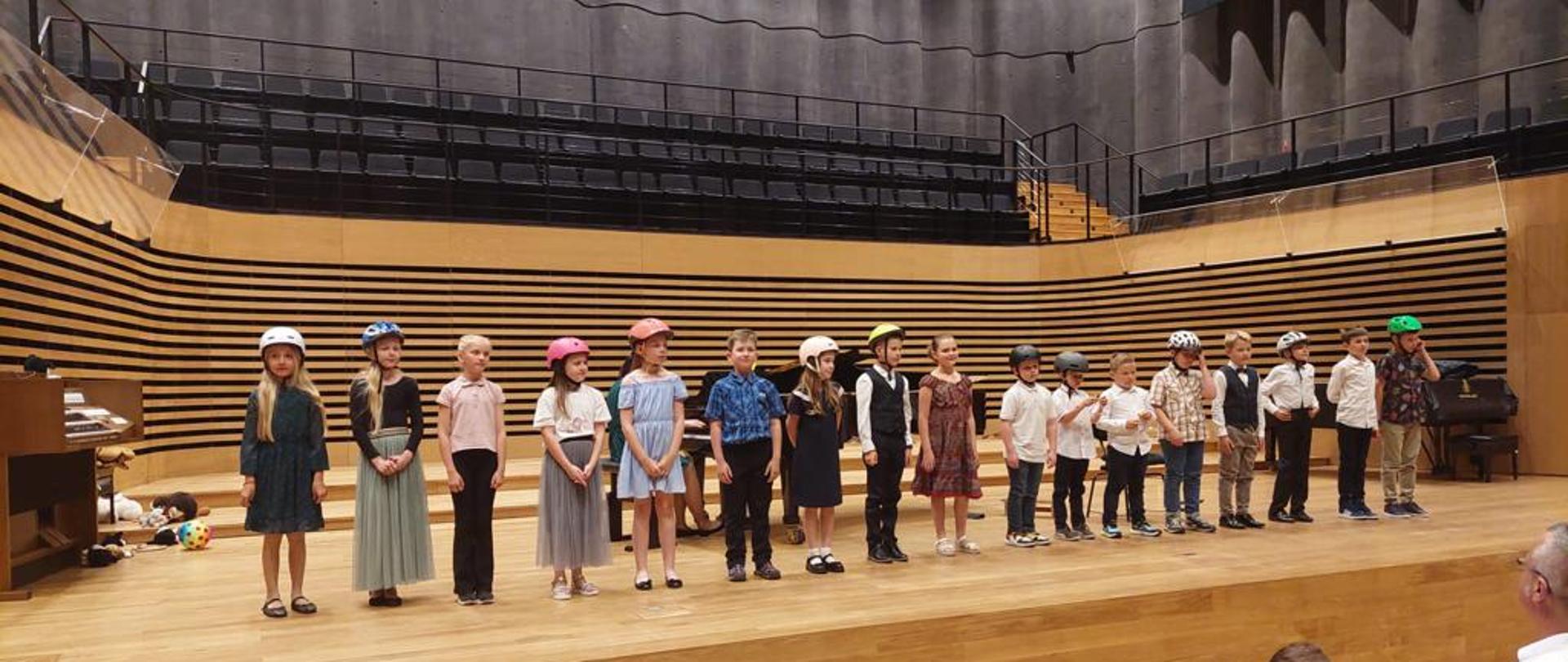 Grupa dzieci (17) stoi w jednym rzędzie na estradzie sali koncertowej. Wszyscy mają założone na głowach kolorowe kaski rowerowe.