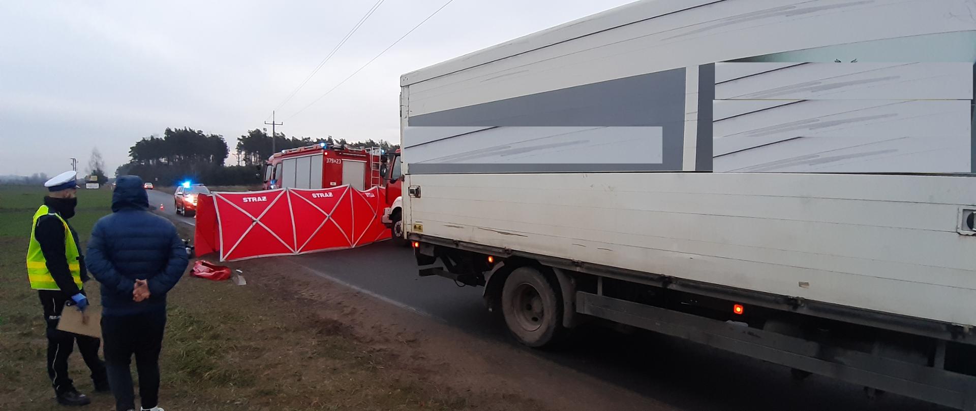 Zdjęcie przedstawia samochód ciężarowy, parawan, samochody ratowniczo - gaśnicze, funkcjonariusza Policji 