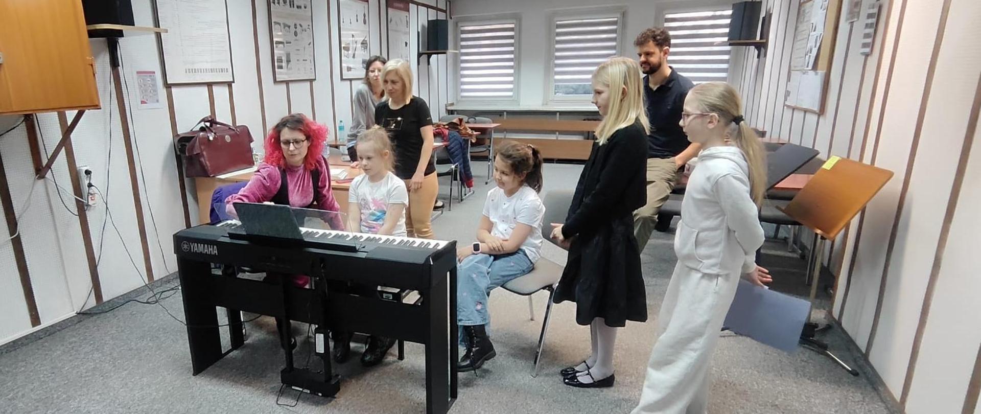 Przy pianinie cyfrowym siedzi dziewczynka i kobieta, dookoła stoją dzieci i nauczyciele i się przypatrują 