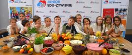Grupa małych dzieci siedzi za stołem, na którym jest dużo kolorowych owoców, za nimi ścianka z napisem Junior Edu-żywienie.