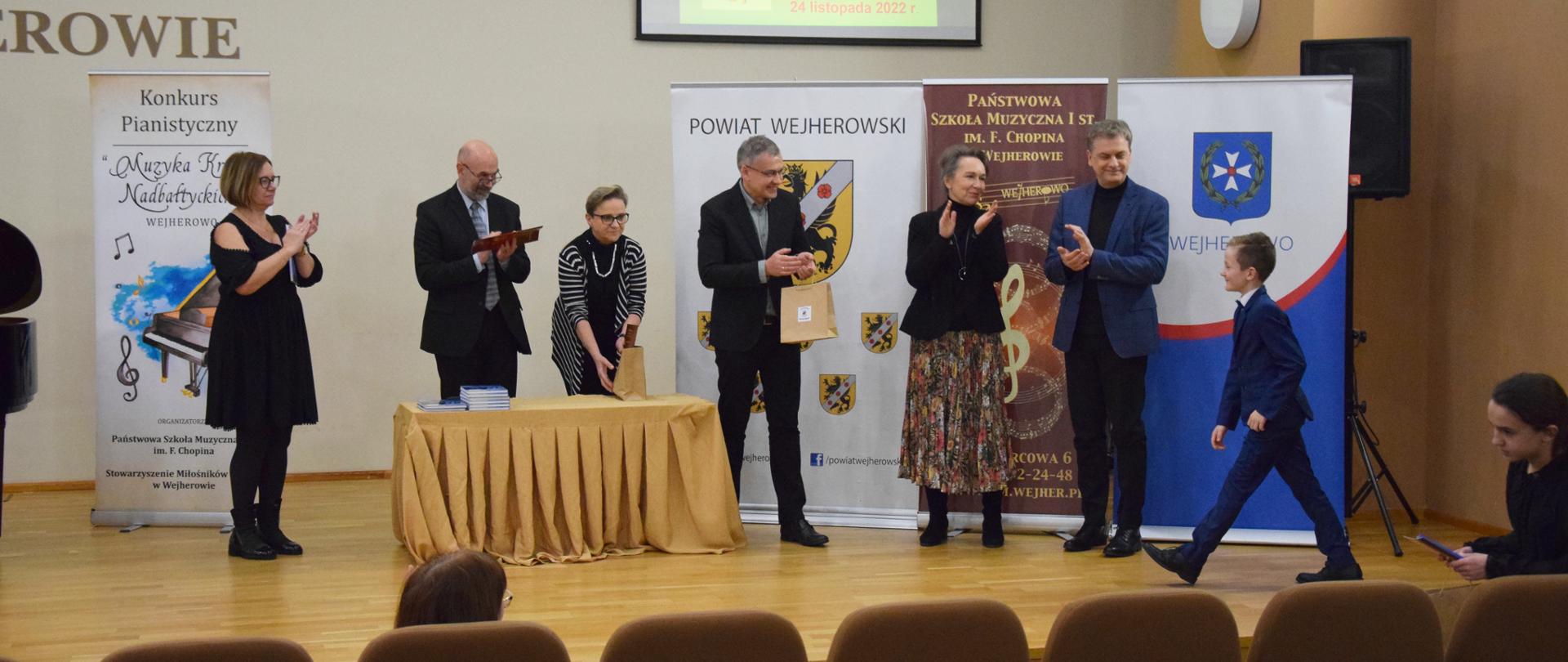 Na zdjęciu widoczna jest stojąca na scenie komisja konkursu pianistycznego oraz laureat Wyróżnienia w grupie II. W tle znajdują się banery konkursowe oraz logo Wejherowa i powiatu wejherowskiego.