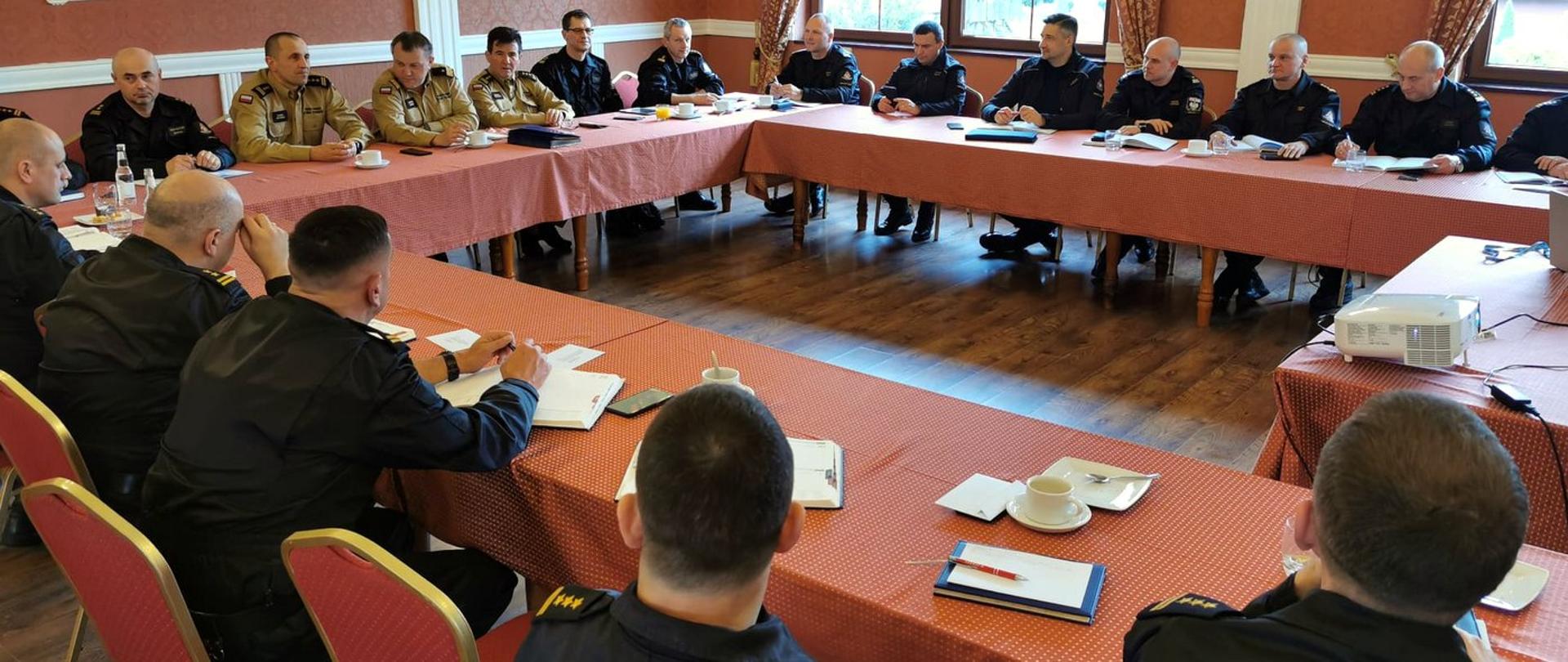 Zdjęcie zrobione w sali szkoleniowej. Przy stołach siedzą oficerowie Państwowej Straży Pożarnej uczestniczący w naradzie. Strażacy ubrani są mundury służbowe.
