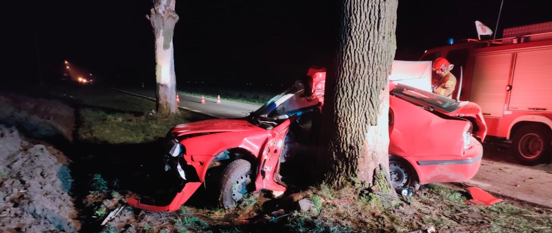 Na zdjęciu widać rozbity czerwony samochód, który stoi przy drzewie. Za samochodem stoi strażak w żółtym stroi i czerwonym hełmie. Za strażakiem stoi czerwony samochód strażacki. W tle widać drzewo, pachołki drogowe, które stoją na drodze. W oddali widać zapalone latarnie uliczne. Jest ciemno, ale miejsce jest oświetlone.