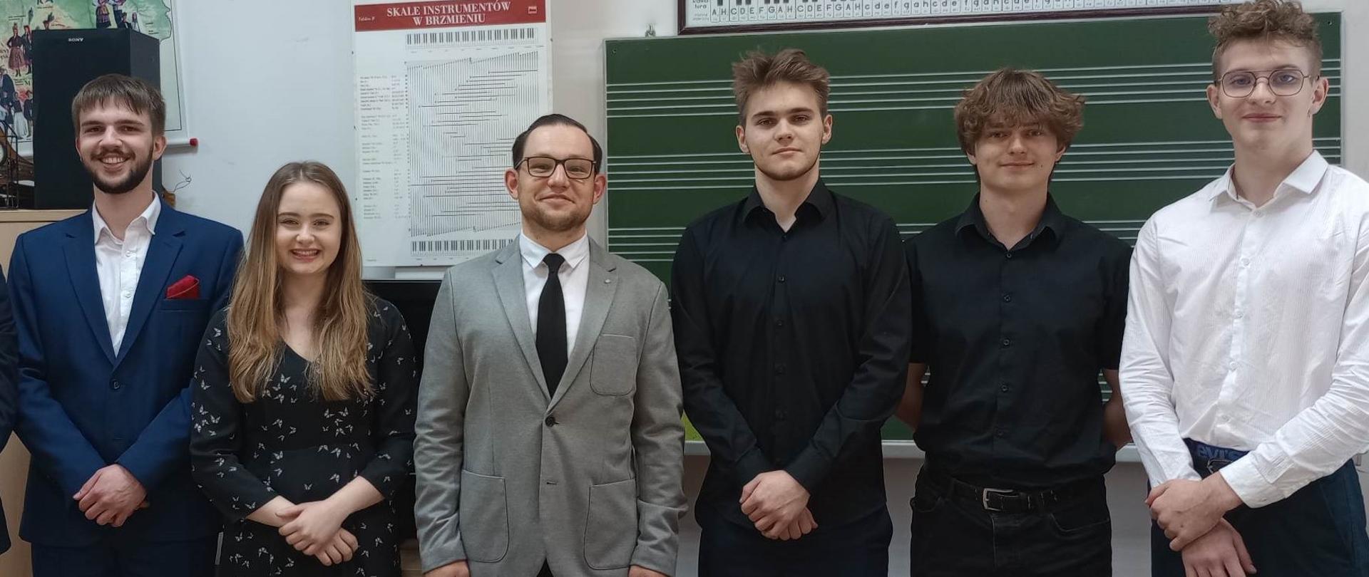 Zdjęcie czterech chłopaków i jednej dziewczyny. Osoby stoją obok siebie, uśmiechają się. W tle tablica w pięciolinie.