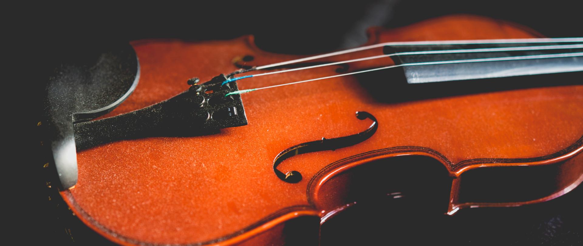 fotografia przedstawia pudło rezonansowe skrzypiec z trzema strunami usytuowane na czarnym tle 