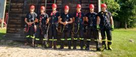 Zdjęcie grupowe uczestników podczas szkolenia z ratownictwa wysokościowego na poziomie podstawowym. Strażacy w umundurowaniu koszarowym, z kaskami na głowach oraz w uprzężach stoją na tle wspinalni na Jednostce Ratowniczo-Gaśniczej w Głuchołazach.