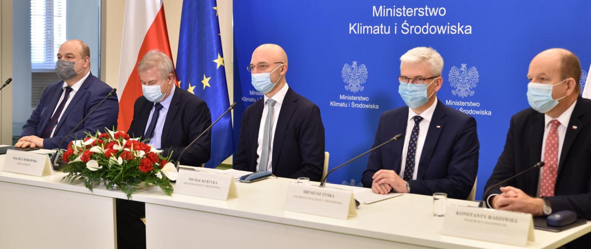 Na zdjęciu znajduje się pięciu elegancko ubranych mężczyzn, którzy siedzą za białym stołem. W tle widać niebieską ściankę z białymi napisami, a po lewej stronie dwie flagi: biało czerwoną oraz granatową z żółtymi gwiazdkami