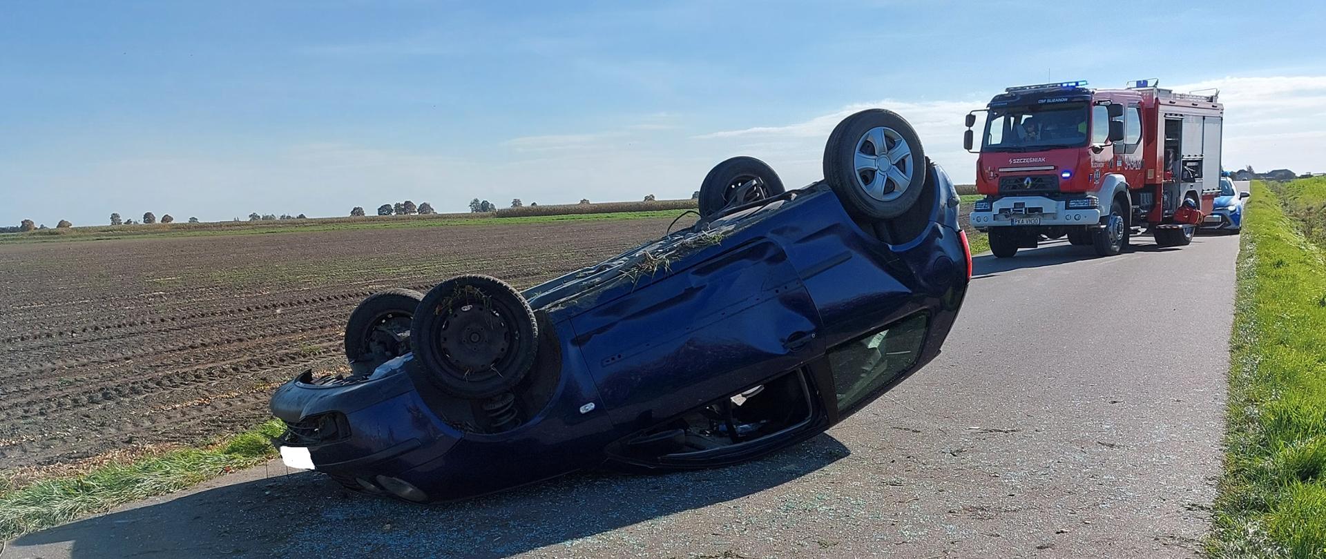 Na zdjęciu uszkodzony pojazd na drodze publicznej. Pojazd leży na dachu. Elementy karoserii na jezdni. Zdjęcie zrobione w ciągu dnia. Na drugim planie fragmenty pól uprawnych. Teren wiejski. 