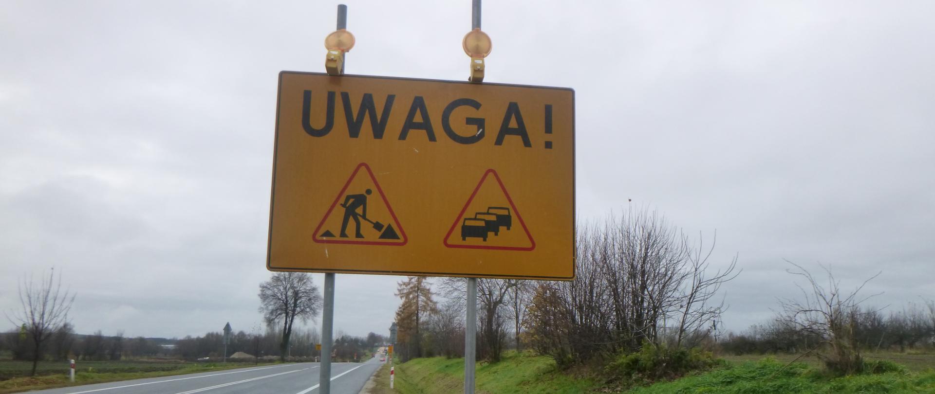 Tablica przy drodze informująca o remoncie i możliwych utrudnieniach w ruchu. Na żółtym tle czarne symbole - drogowiec z łopatą i samochody stojące w korku