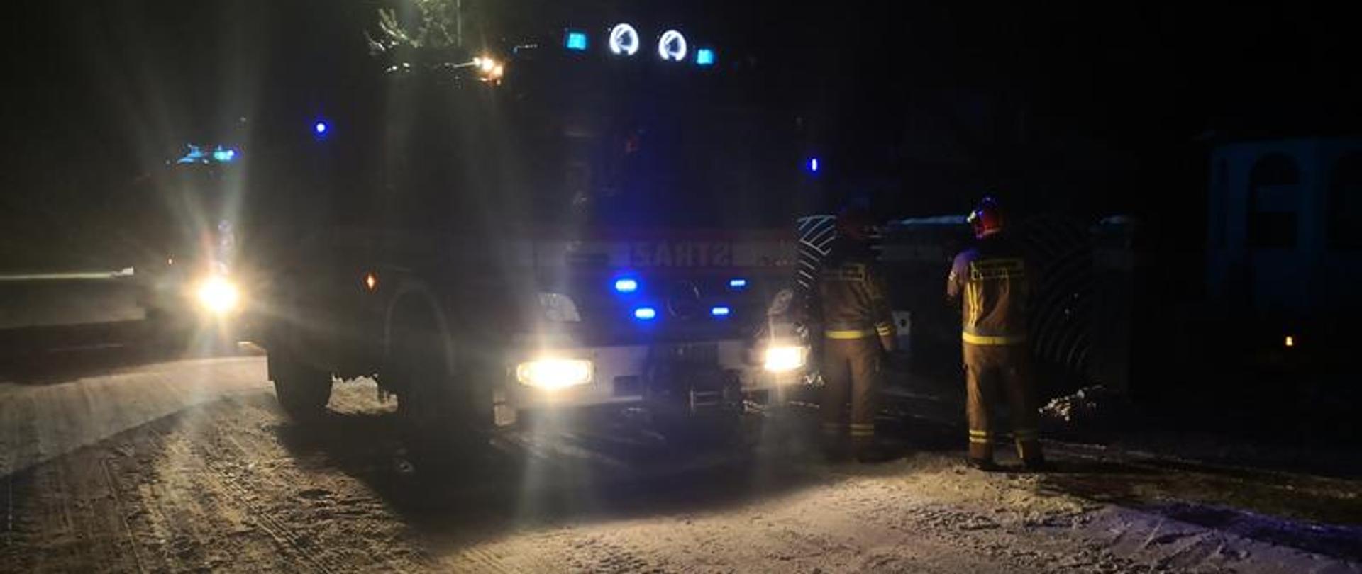Dwóch strażaków ubranych w środki ochrony osobistej prowadzą działania związane z pożarem sadzy w przewodzie kominowym. Obok posesji ustawione są pojazdy pożarnicze z włączoną sygnalizacją świetlną.